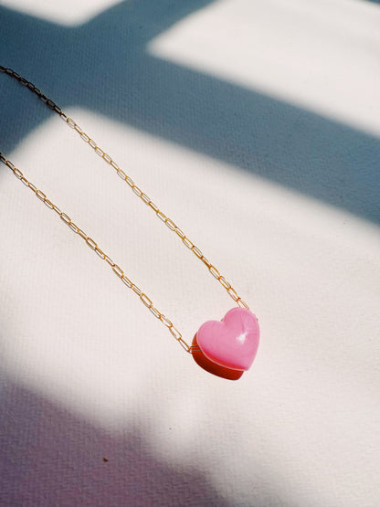 The Acrylic Heart Necklace, featuring a gold filled chain and pink acrylic heart pendant, rests on a textured white surface with sunlight casting soft shadows over this elegant jewelry piece.