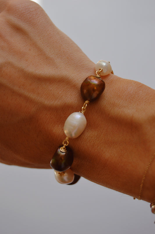 A close-up of a wrist wearing the Chocolate and White Pearl Bracelet, showcasing alternating white and brown cultured freshwater pearls on a 14k gold-filled chain against a neutral backdrop.