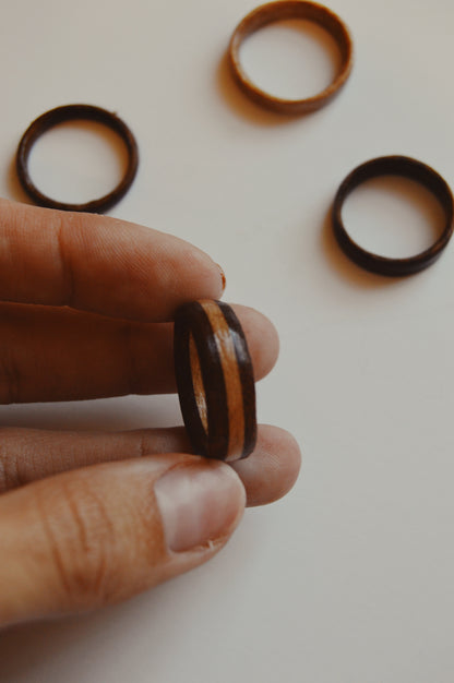 A hand displays the Wooden Ring- Striped, an artisan piece with a central white oak stripe, while three more handmade Wooden Ring- Striped pieces rest on a white surface in the background.