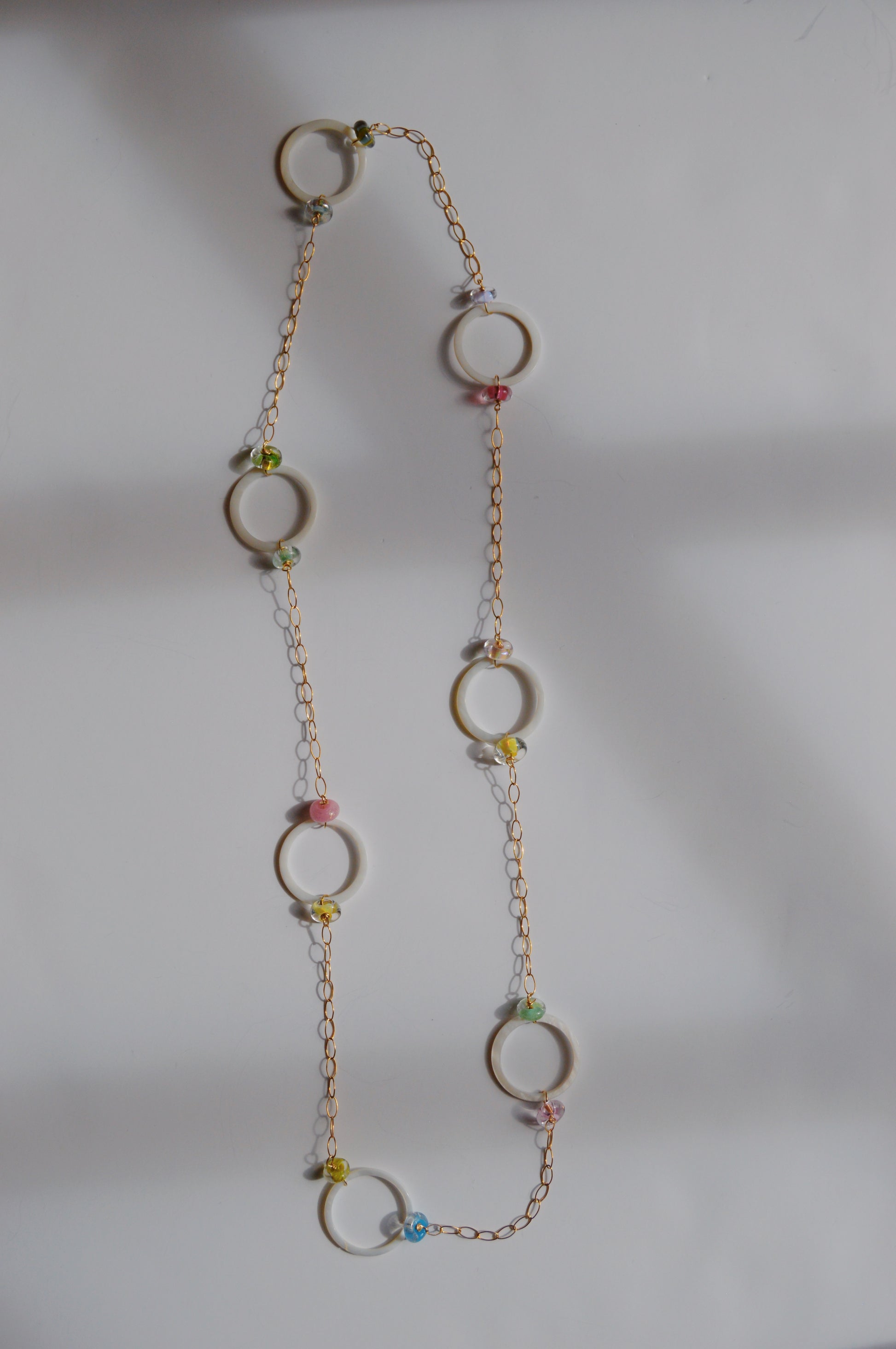 The Long Glass and Circle Layer necklace showcases large gold rings linked by a delicate 14k gold-filled chain and accented with colorful handmade lampwork glass beads, all displayed on a white background.