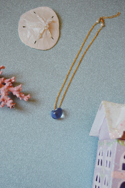 The Grand Bubble Single - Corduroy necklace with a blue pendant is displayed on a glittery surface, accompanied by a sand dollar, freshwater pearls, pink coral, and the roof of a decorative pastel house.