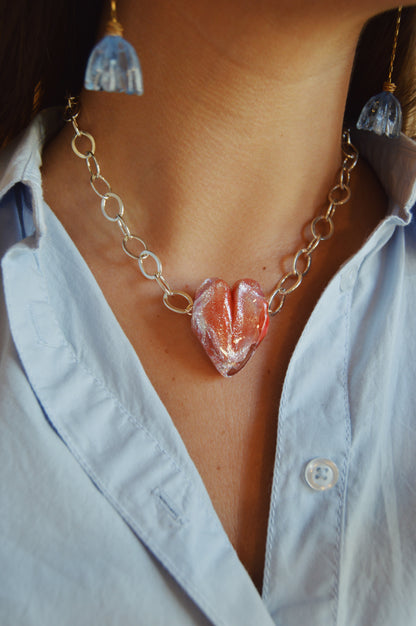 A close-up of a person wearing a light blue button-up shirt, blue flower earrings, and the Chunky Sterling Silver Glass Heart Necklace with a handblown glass heart pendant crafted by local glass artists.