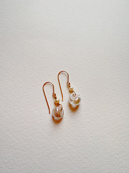 The Bubble Minis are delicate drop earrings with gold-filled findings, each adorned with a small white bead and a clear round glass bead, displayed on a textured white surface.