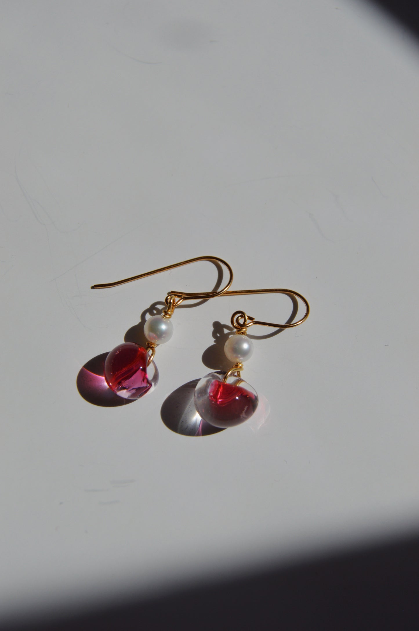 Bubble Minis: Gold hook earrings featuring freshwater pearls, translucent pink teardrop charms, and delicate glass bead accents, resting on a white surface with a shadow at the corner.