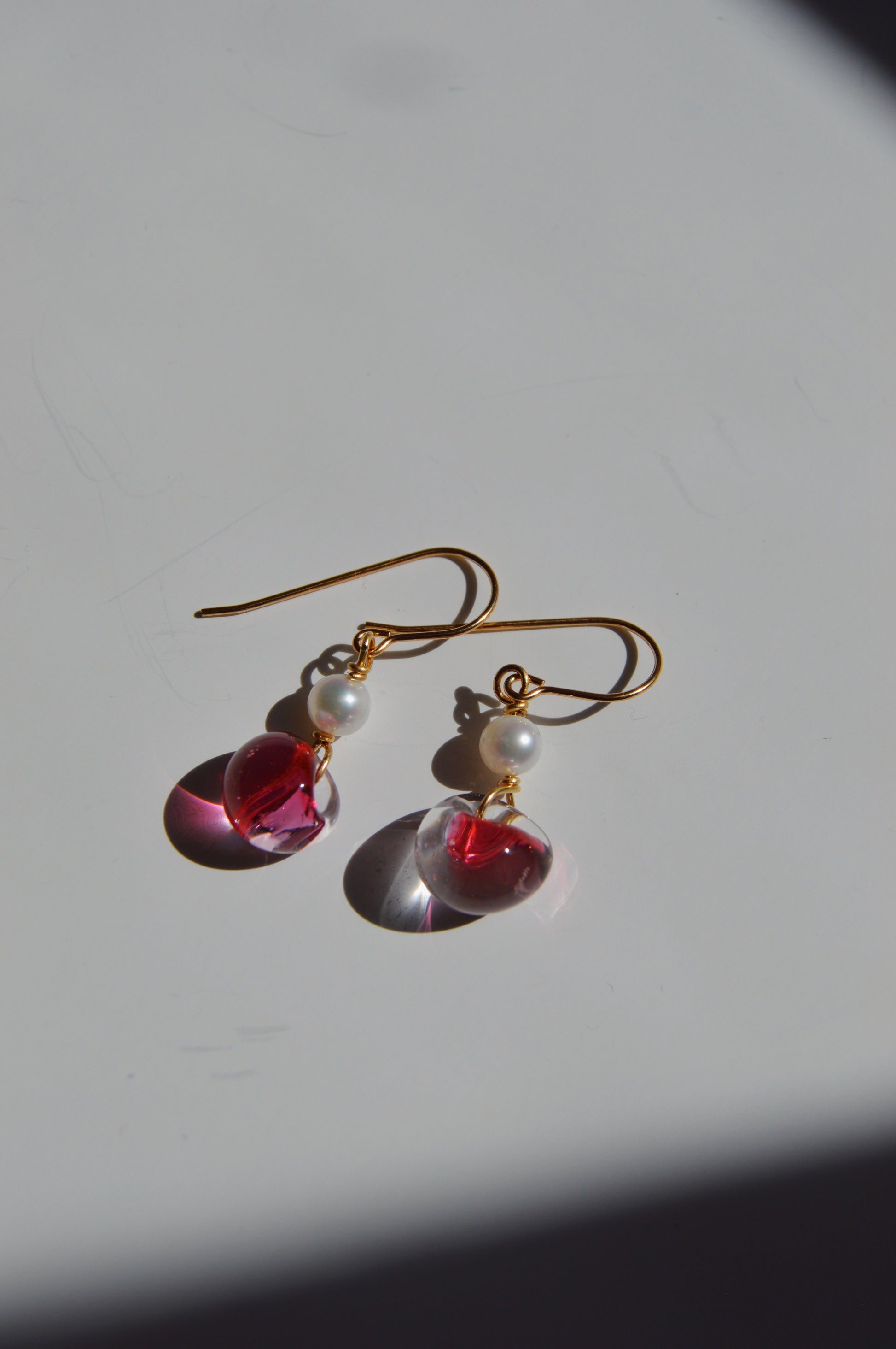Bubble Minis: Gold hook earrings featuring freshwater pearls, translucent pink teardrop charms, and delicate glass bead accents, resting on a white surface with a shadow at the corner.