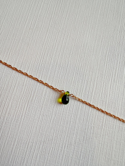 The Dewdrop Bracelet, featuring a 14k gold filled adjustable chain and a small green glass teardrop charm, rests on a white textured surface.
