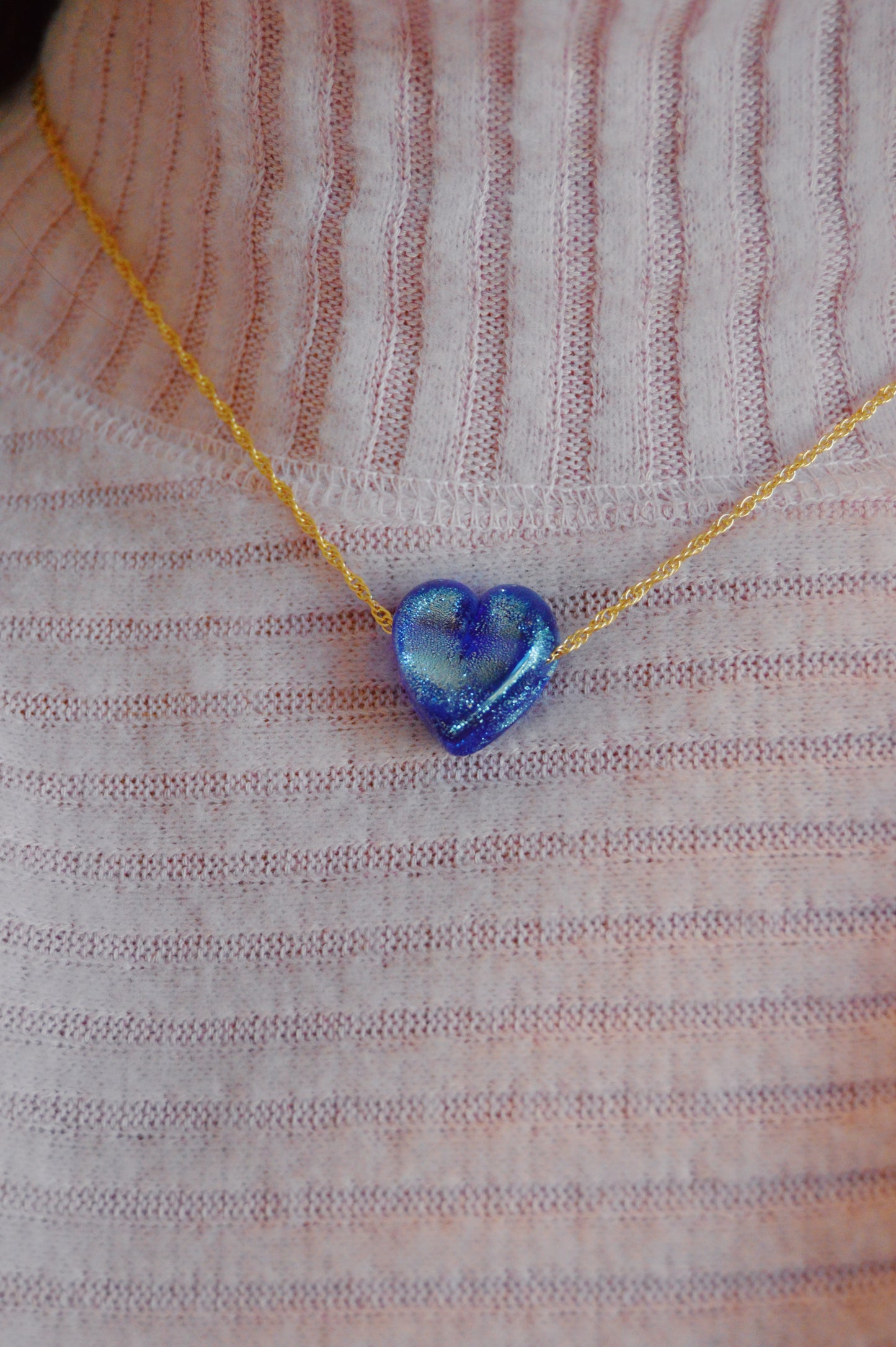 A person wearing a ribbed light pink sweater and a gold-filled chain necklace featuring an artisan touch: a shiny, handmade lampwork blue Glass Heart Necklace.