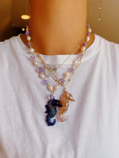 A person models the Glass Seahorse Layer: one necklace with cultured freshwater pearls and a blue glass seahorse pendant, another with 14k gold filled links and a clear glass seahorse pendant, both worn over a plain white t-shirt.