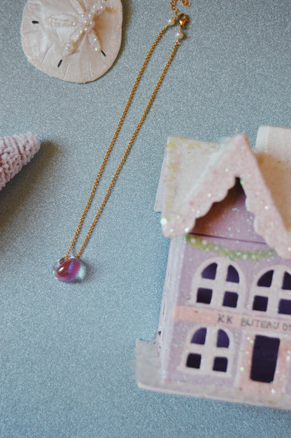 The Grand Bubble Single - Shimmer Pink, a gold necklace with a purple pendant and 14k gold filled accents, rests on a sparkly surface beside a pastel pink ceramic house, sand dollar, and seashell.