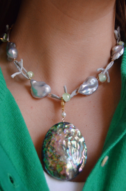 A close-up shows a person wearing the Abalone Charm necklace, featuring large irregular freshwater pearls, small round beads, and a bold abalone shell pendant with gold-filled accents that complement their green top.