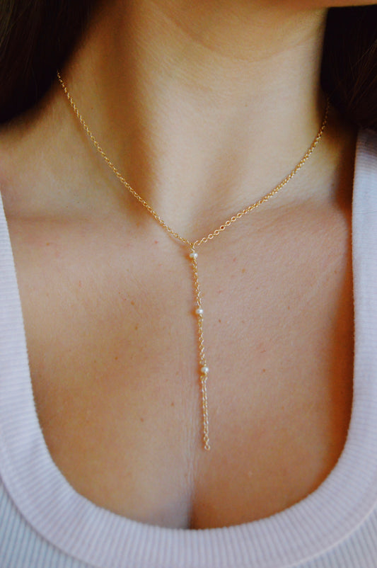 A close-up of a person wearing the Dainty Pearl Lariat, a gold Y-shaped necklace with small pearls on a 14k gold filled chain, paired with a white ribbed tank top. The necklace sits elegantly above the chest.