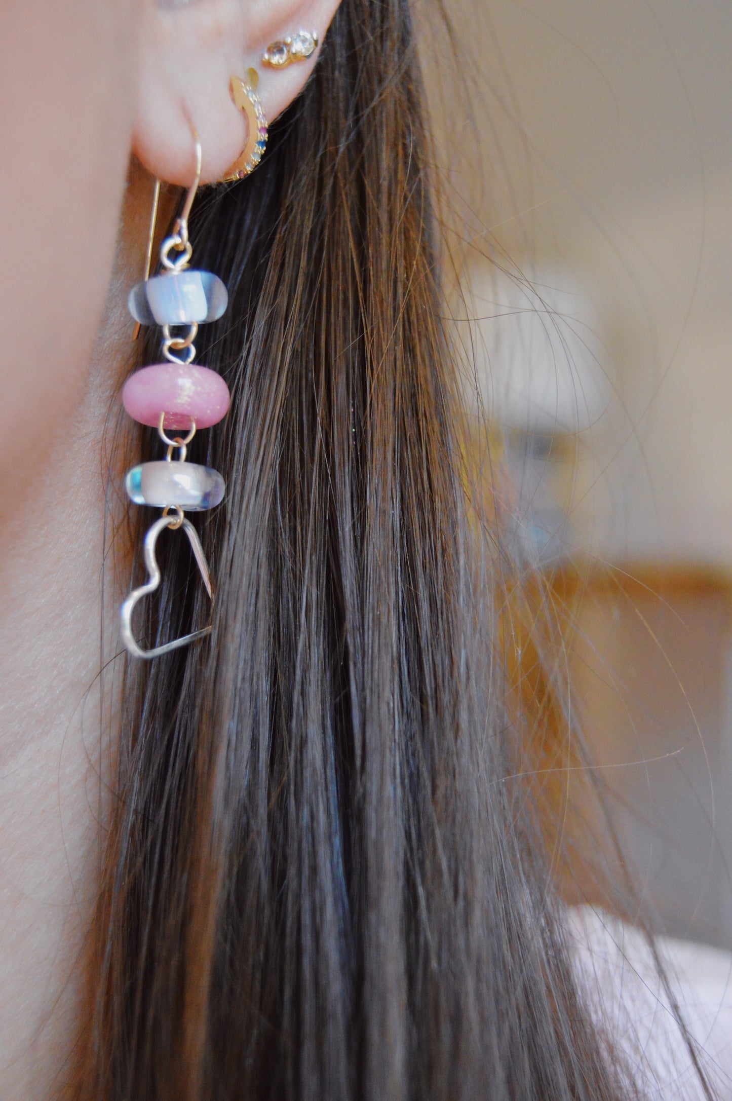 Close-up of a person's ear adorned with handcrafted jewelry: multiple gold earrings and Silver Heart Drops, featuring lampwork glass beads and a sterling silver heart-shaped wire. The person's straight brown hair is visible in the background.