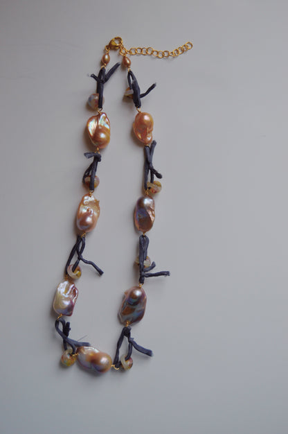 The Silk and Pearl Layer- Blueberry Puka necklace features irregular metallic freshwater pearls on black knotted cords with a gold adjustable clasp, displayed on a light gray background.