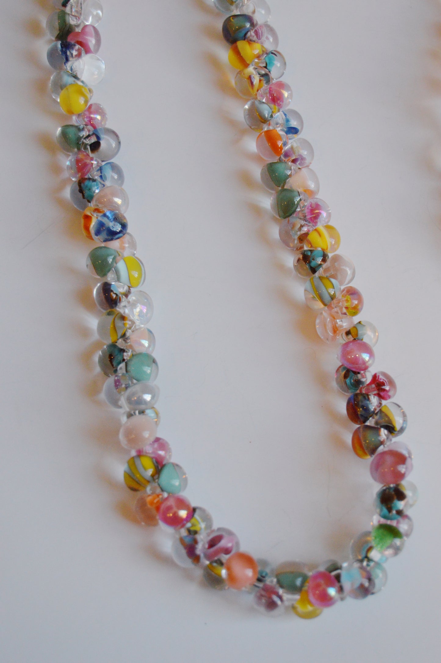 The Rainbow Bubble Glass Necklace, a handmade piece with round lampwork glass beads in pastel pink, blue, yellow, and green, is arranged in a loose curve on a white surface.