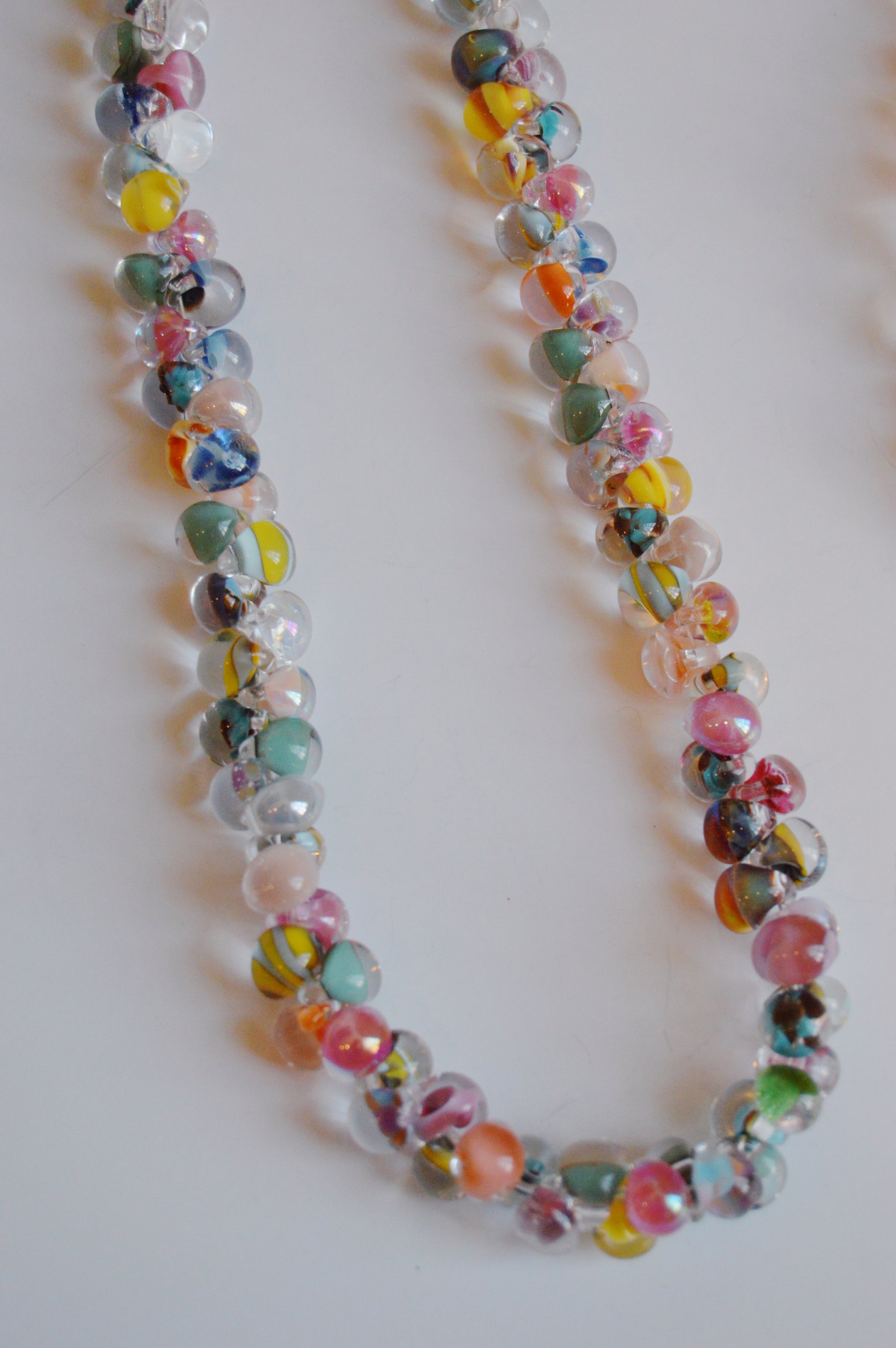 The Rainbow Bubble Glass Necklace, a handmade piece with round lampwork glass beads in pastel pink, blue, yellow, and green, is arranged in a loose curve on a white surface.