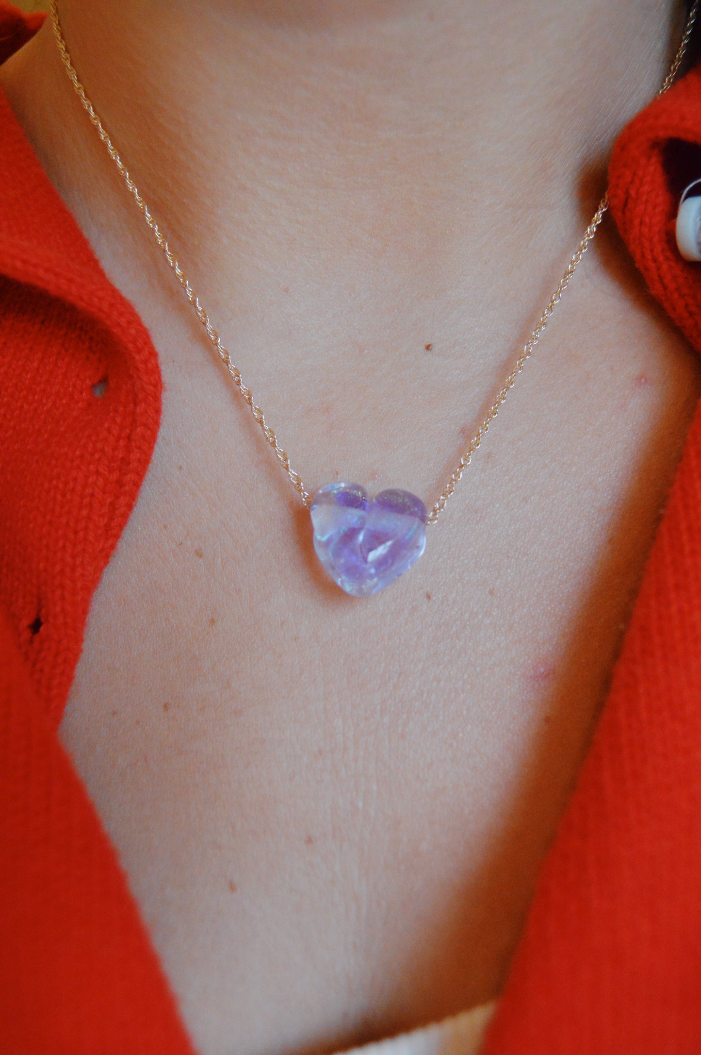 A close-up of a person wearing the Glass Heart Necklaces- Purples, featuring a gold-filled chain and a handmade purple lampwork glass heart pendant, set against bare skin and a red open-collar shirt—an elegant artisan piece.