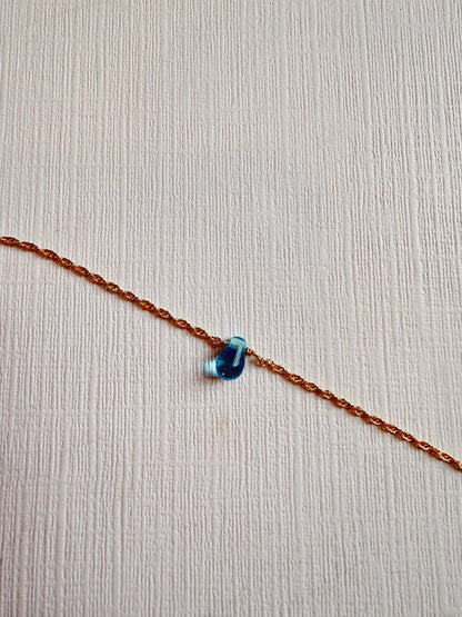 The Dewdrop Bracelet, featuring a delicate 14k gold filled chain and a small blue teardrop-shaped pendant, is displayed diagonally on a textured white surface.