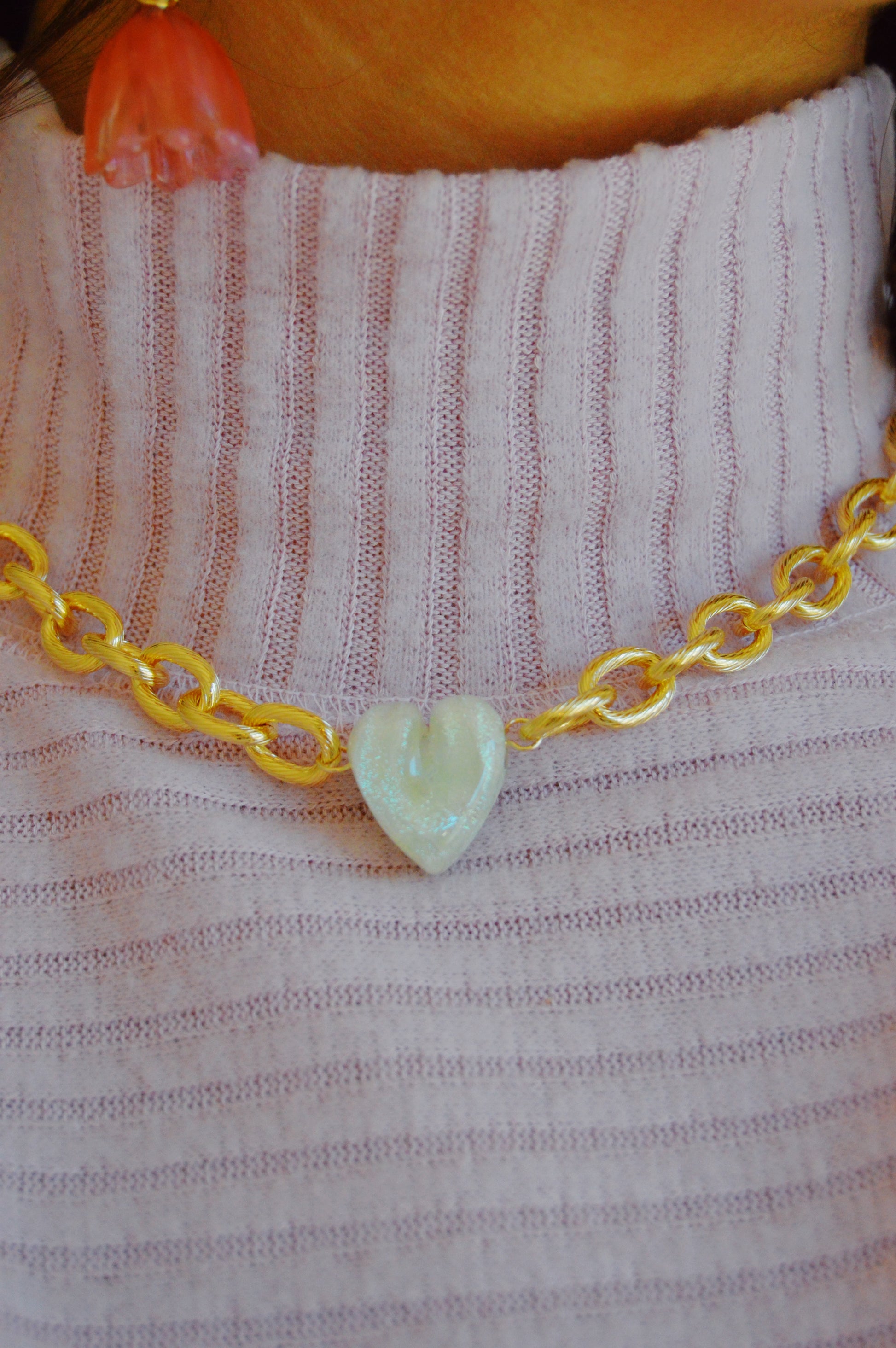 A close-up of the Chunky Plated Glass Heart Necklace—Whites & Clears, featuring a gold plated chunky chain with a handmade glass heart pendant, worn over a light purple ribbed top with part of a pink flower-shaped earring showing.