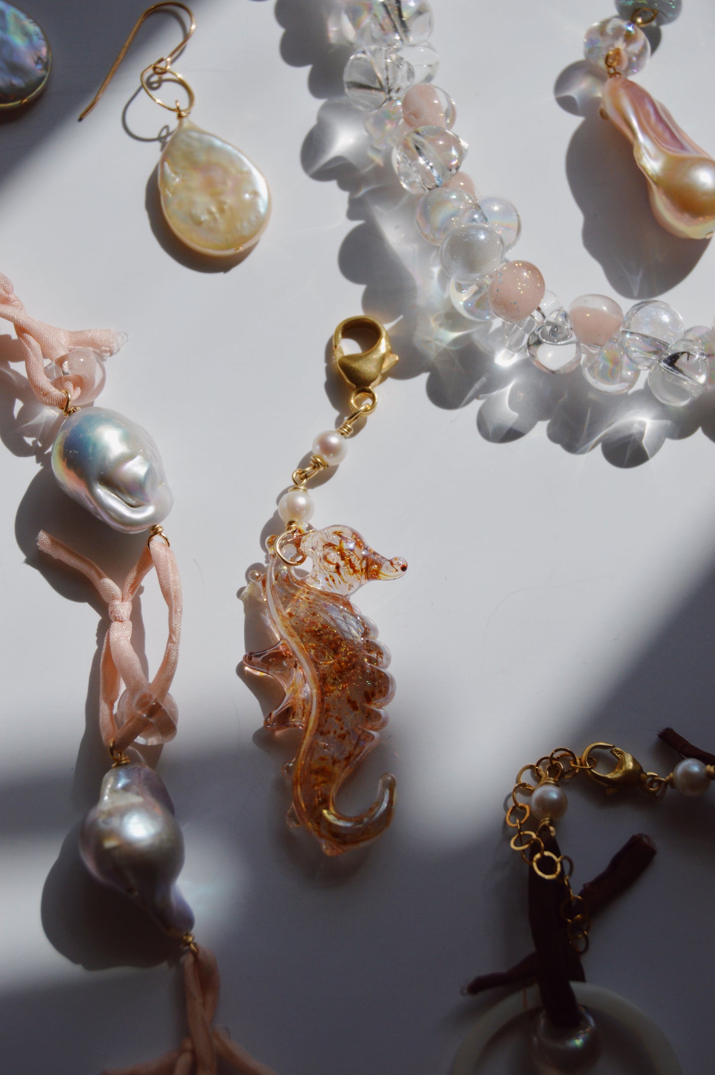 A close-up of various jewelry on a white surface features a Seahorse Charm, freshwater pearl earrings, and beaded bracelets with 14k gold-filled clasps, all illuminated by natural sunlight.
