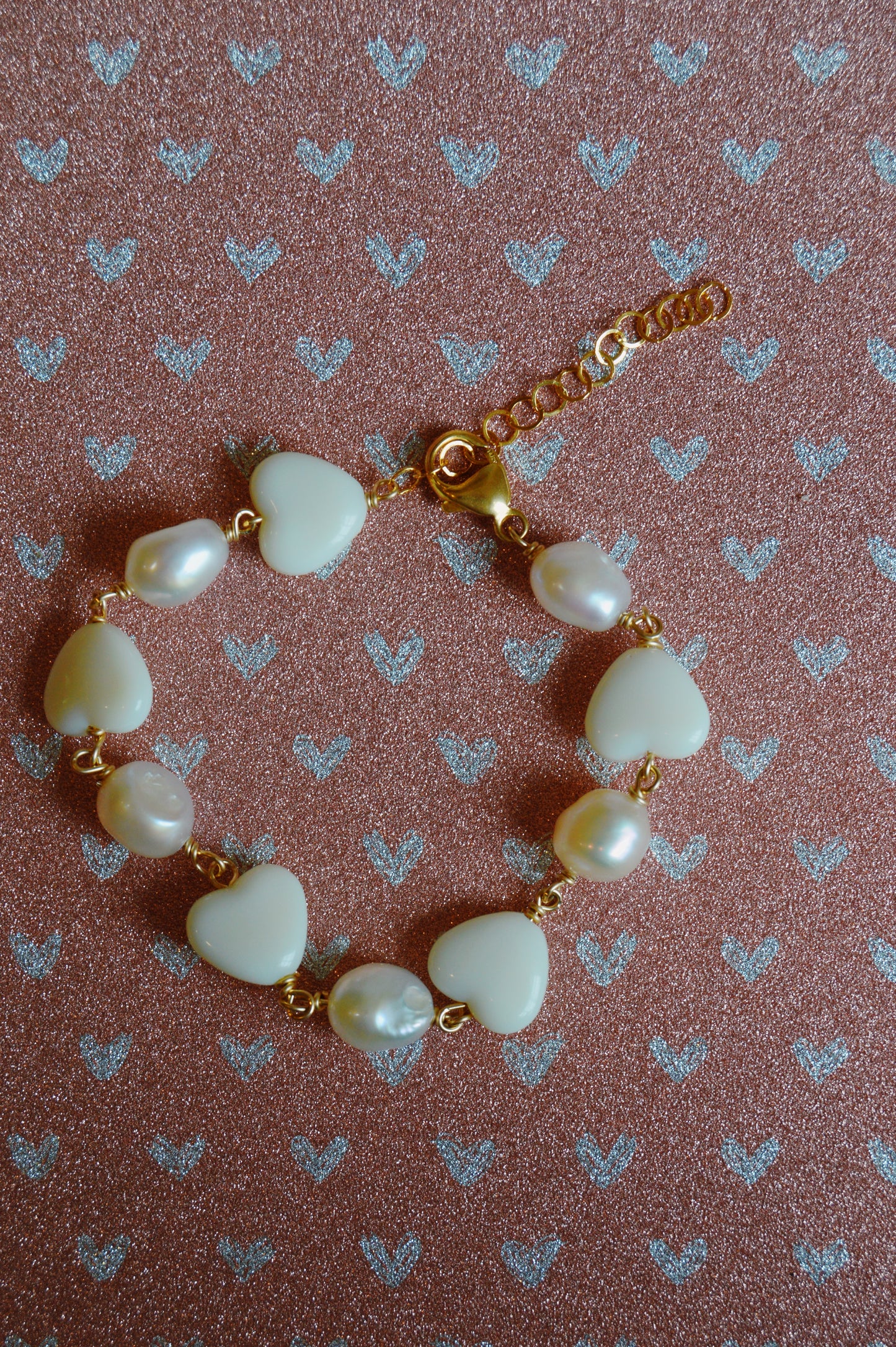 The Candy Bracelet-White features alternating white heart-shaped and round beads on a gold chain, displayed on a pink surface with a glittery silver heart pattern.