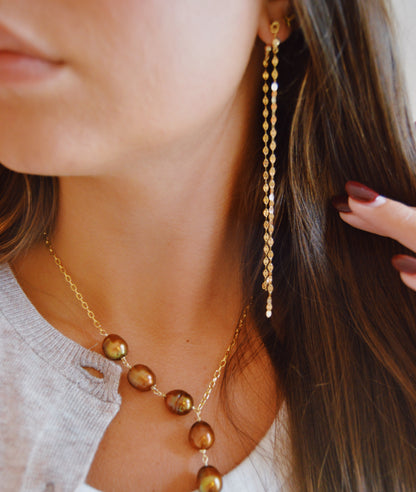 A woman with long brown hair and manicured nails wears Sway Studs—long, dangling 14k gold filled earrings—and a gold chain necklace with brown pearls. She’s dressed in a white top topped with a light cardigan.