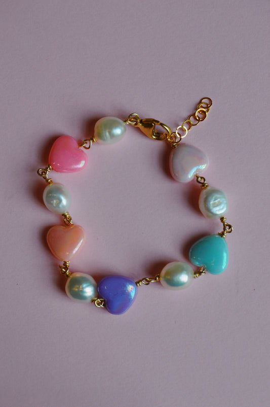 The Candy Bracelet features a gold chain with alternating pastel heart-shaped beads and round pearl-like beads, shown on a light pink background.
