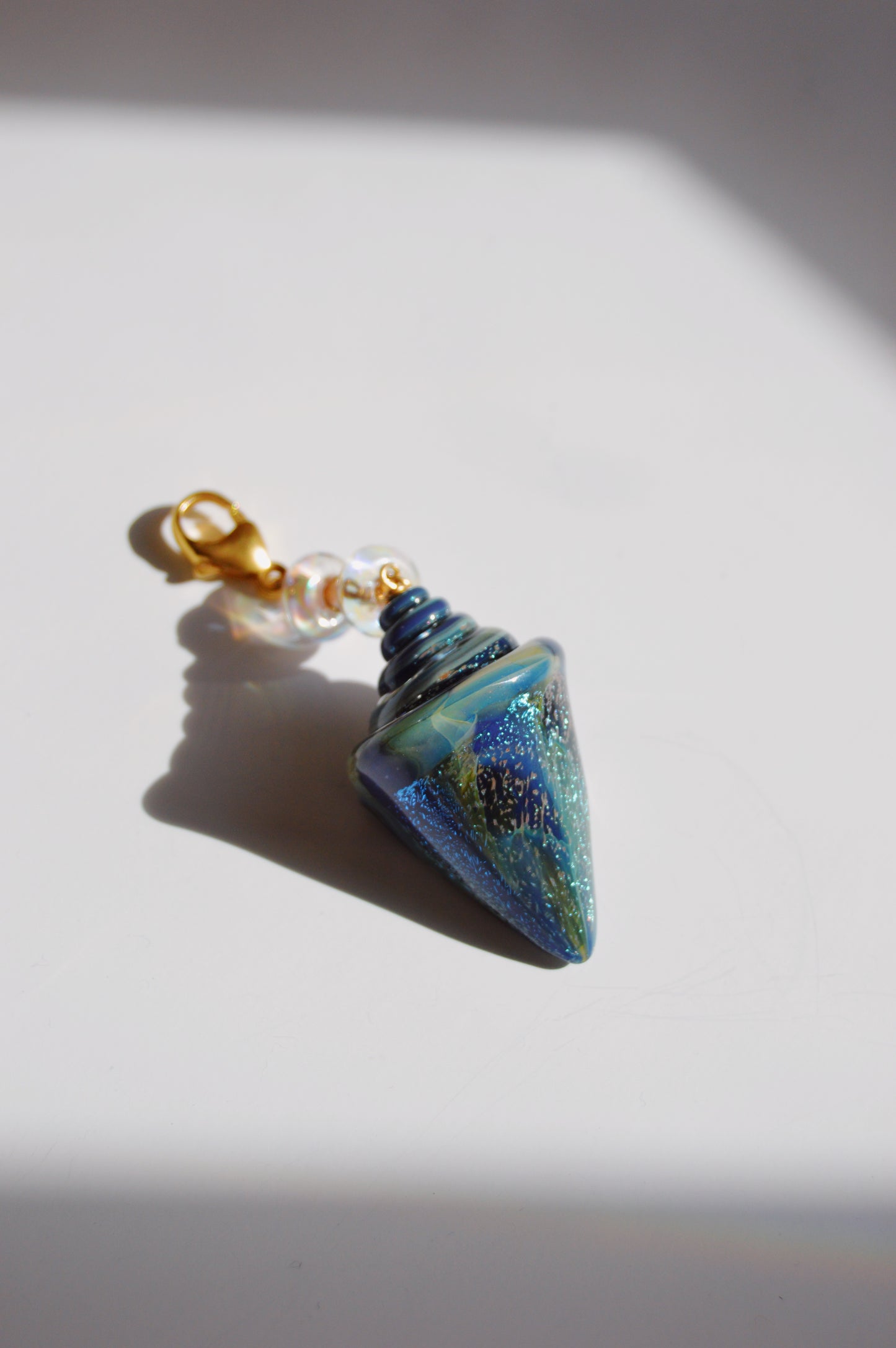 The Glass Shell Charm, an iridescent blue and green lampwork glass pendant with a 14k gold-filled clasp and pearl-like beads, casts a soft shadow on a white surface.