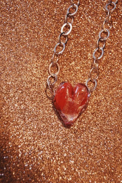 The Chunky Sterling Silver Glass Heart Necklace, featuring a red handblown glass heart pendant made by local artists, is showcased on a glittery gold surface.