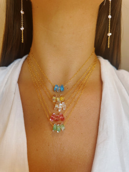 A woman wears the Dainty Donut Layer—layered 14K gold chain necklaces with colorful glass donut bead pendants—and long gold pearl earrings, shown from the chest up in a white top.