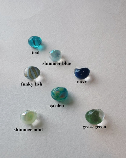 Seven Bubble Drops—handblown glass beads in teal, shimmer blue, funky fish, navy, garden, shimmer mint, and grass green—are arranged on a white surface.