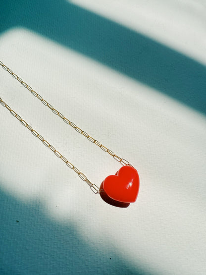 The Acrylic Heart Necklace features a bright red acrylic heart pendant on a 14k gold filled chain, displayed on a textured white surface with sunlight and shadows forming bold diagonal stripes in the background.