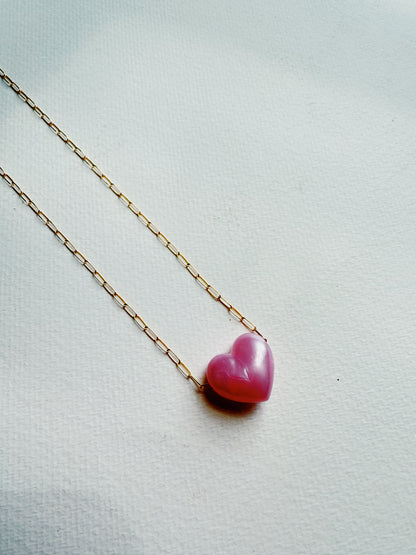The Acrylic Heart Necklace features a pink acrylic heart pendant on a 14k gold filled chain, elegantly displayed on a white textured surface.