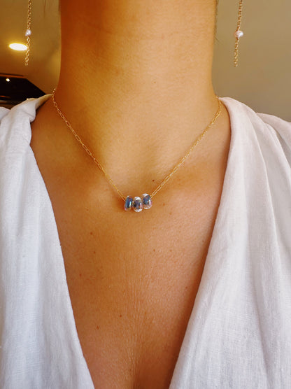 A person wears the Dainty Donut Layer—a delicate 14K gold chain necklace with three small blue gemstone beads and a secure gold-filled clasp—paired with a white V-neck top. Matching gold earrings with pearl drops are also partially visible.