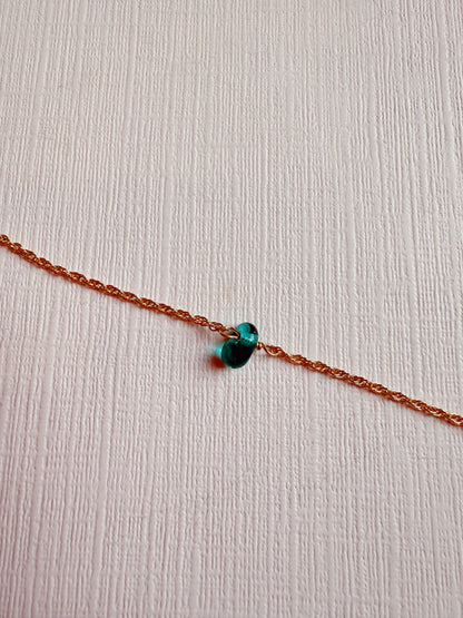 The Dewdrop Bracelet, featuring a 14k gold-filled chain and a small turquoise-green glass bead, rests on a textured white surface.