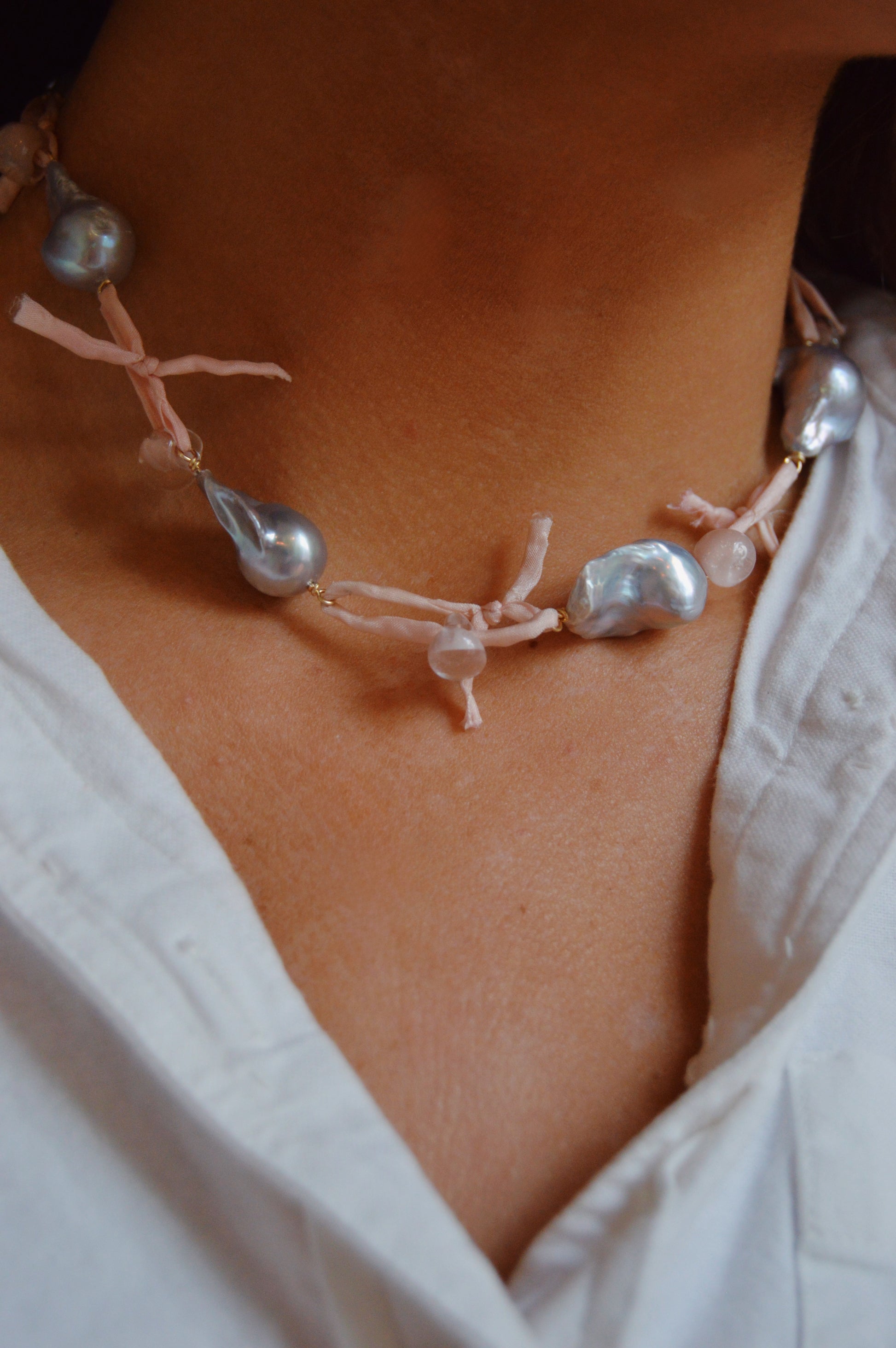 Close-up of a person wearing the Silk and Pearl Layer- Blossom necklace, featuring large irregular freshwater pearls and light pink fabric knots, paired with a white shirt.