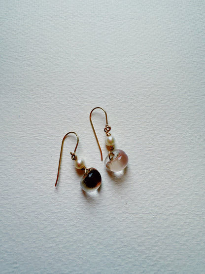 The Bubble Minis are delicate drop earrings with gold hooks, each adorned with a small freshwater pearl and a translucent glass bead, showcased on a textured white surface.