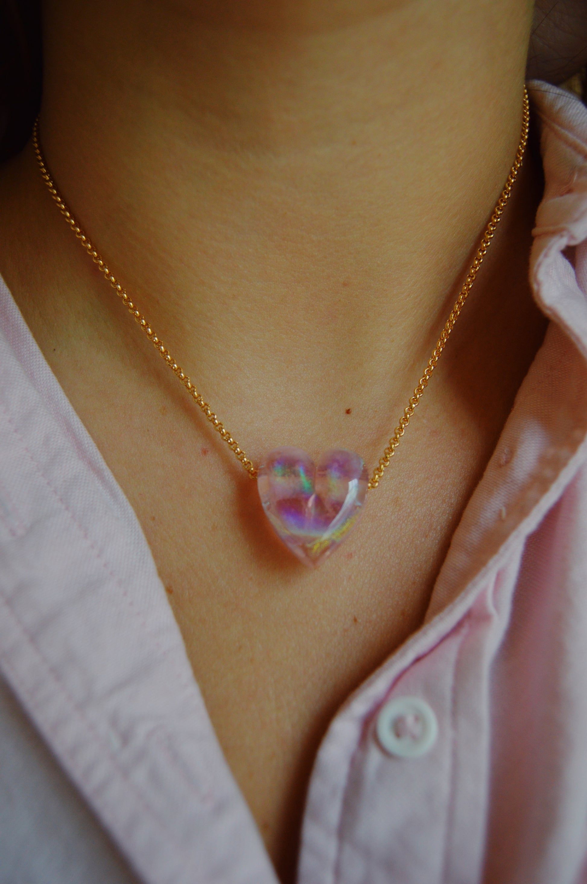 A close-up of someone wearing a Glass Heart Necklaces- Pinks: a 14k gold-filled chain with a handmade lampwork glass heart pendant, shimmering against their light pink collared shirt.