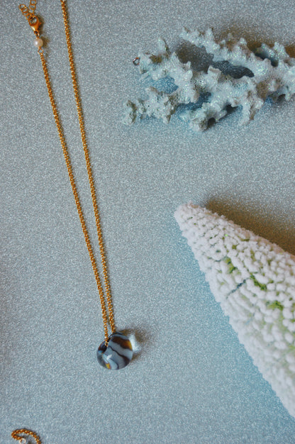 The Grand Bubble Single - Funky Fish gold chain necklace, featuring a round striped pendant and handblown glass beads, rests on a glittery light blue surface with two white coral decorations.
