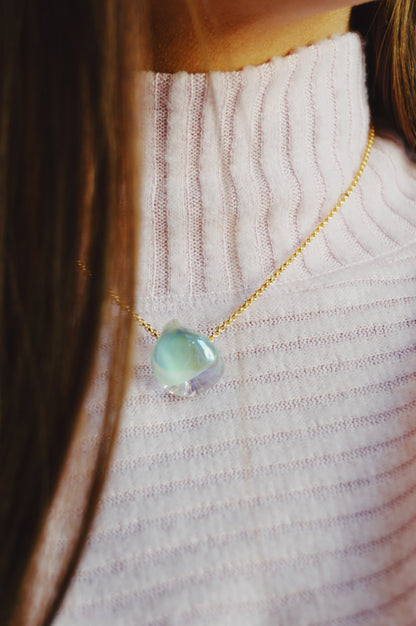 A close-up of a person wearing a ribbed, light pink turtleneck and brown hair, styled with the Grand Bubble Single - Shimmer Blue: a 14k gold filled chain necklace featuring an iridescent stone pendant.