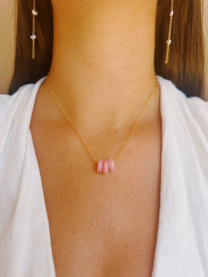 A woman in a white top showcases the Dainty Donut Layer: a delicate 14K gold chain necklace with two pink glass donut beads, paired with matching gold earrings featuring pearl-like accents.