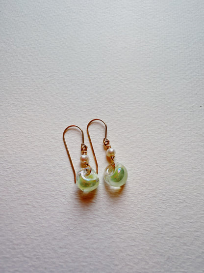 The Bubble Minis are delicate drop earrings with gold filled findings and hooks, each showcasing a small white freshwater pearl and a translucent pale green glass bead, displayed on a textured white background.