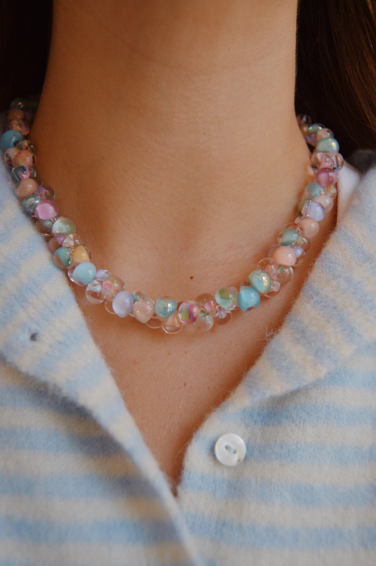 A close-up of a person wearing the Bubble Glass Necklace- Spring Mix, a pastel handmade necklace with pink, blue, and iridescent beads. The person is dressed in a blue and white striped button-up shirt.