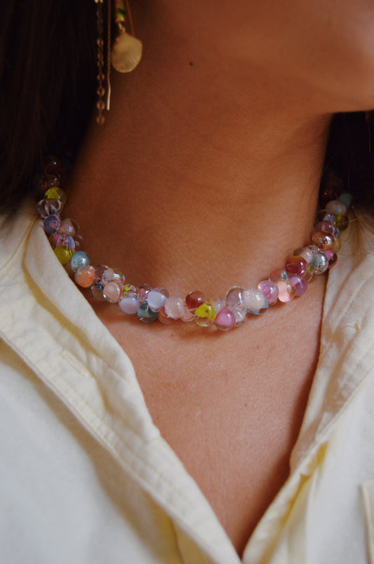 Close-up of a person wearing the Rainbow Bubble Glass Necklace featuring pastel lampwork beads, paired with a light cream collared shirt; an earring is partially visible, and short brown hair frames their neck.