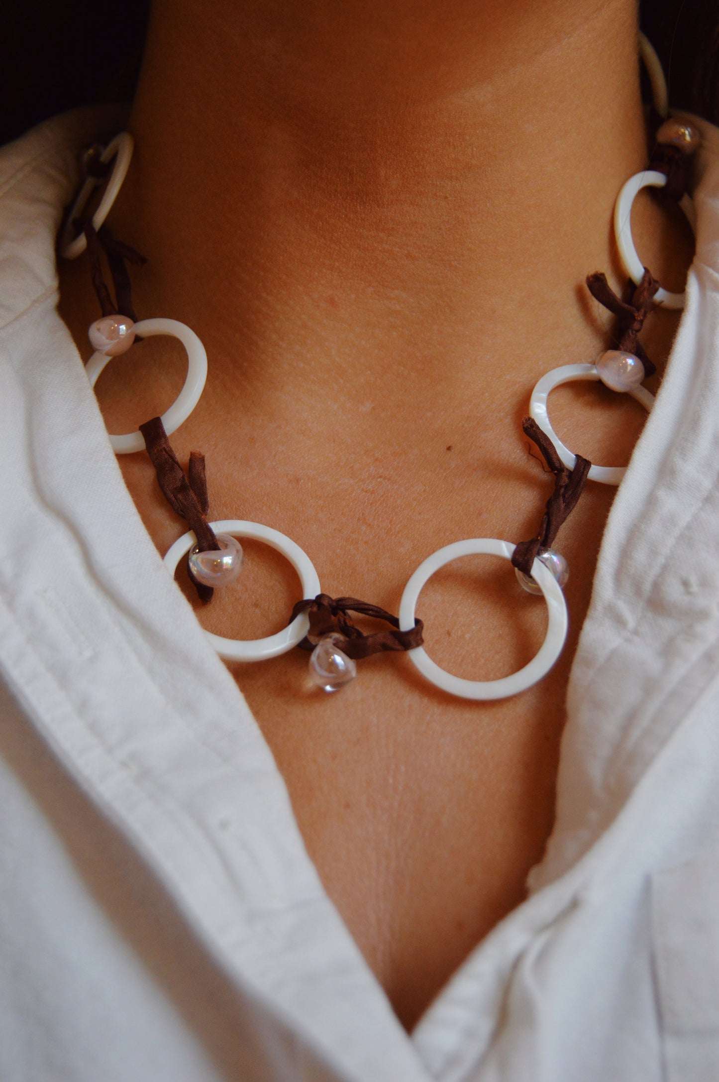 A close-up of a person wearing a white collared shirt and the Silk and Circle Layer—a handmade necklace of large white rings, brown cord, and small translucent lampwork glass beads, creating an elegant look.
