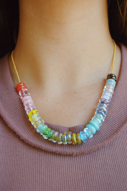 A close-up of a person wearing the Rainbow Lots of Donuts 14k gold filled chain necklace with colorful round handblown glass beads, resting on their neck above a mauve ribbed shirt.