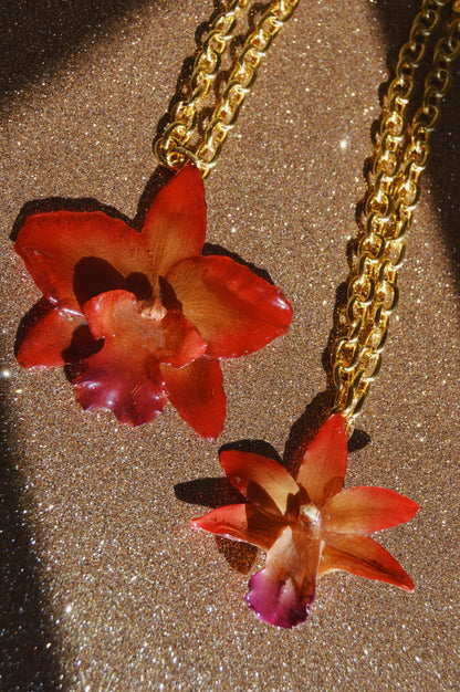 Two Large Orchid Necklaces, each featuring a preserved orchid pendant in red and purple, are displayed on a glittery brown surface as sunlight creates captivating shadows.