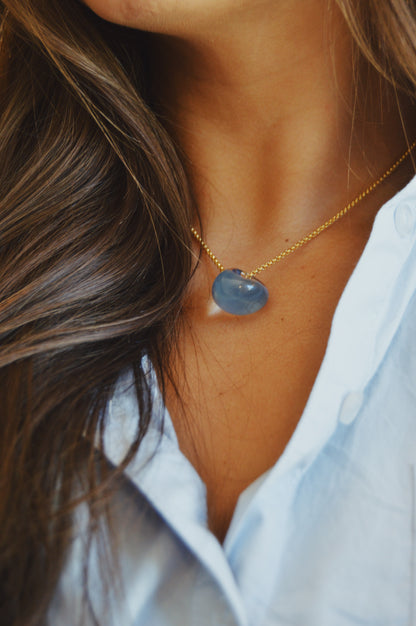 A close-up of a woman in a light blue shirt wearing the Grand Bubble Single - Corduroy necklace, featuring a gold-filled chain with a blue stone pendant. Only her neck, collarbone, and part of her shirt are visible.