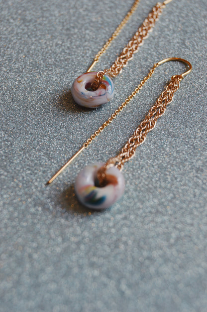 The Glass Donut Threaders are gold chain dangle earrings featuring small, marbled purple and blue handblown glass beads on a 14k gold filled chain, shown on a sparkling silver surface.