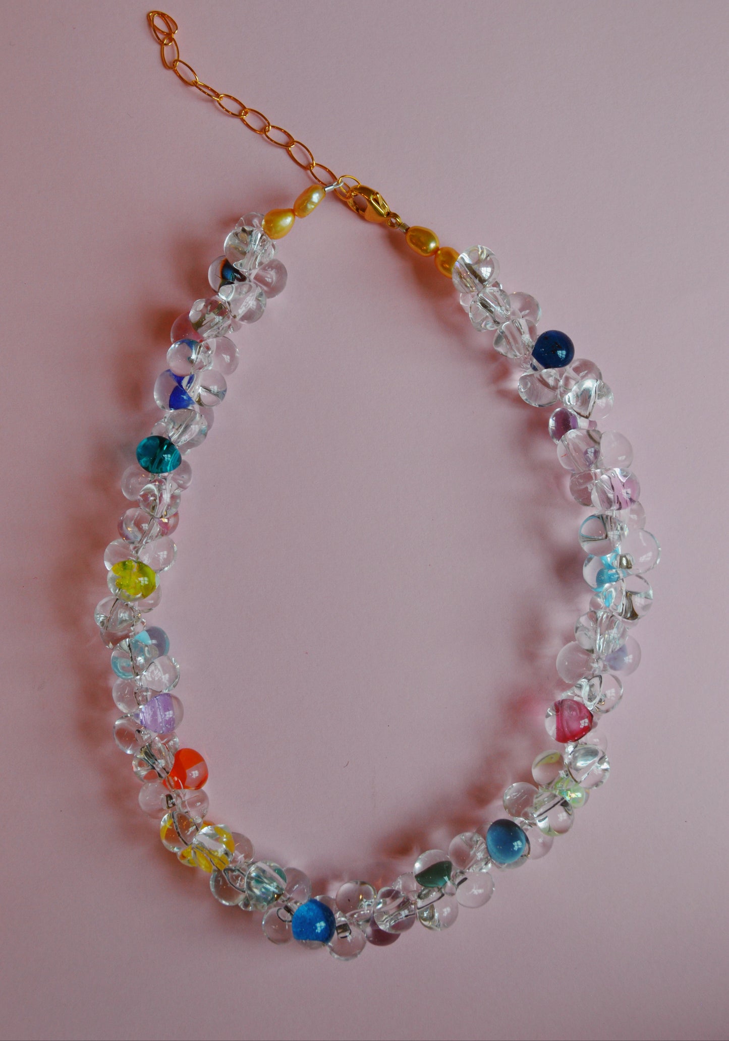 The Valentine Bubble Glass Necklace- Sprinkles features clear and colorful round beads—blue, yellow, red, and green—on a gold chain with clasp, displayed against a light pink background.