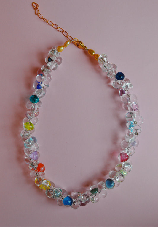 The Valentine Bubble Glass Necklace- Sprinkles features clear and colorful round beads—blue, yellow, red, and green—on a gold chain with clasp, displayed against a light pink background.